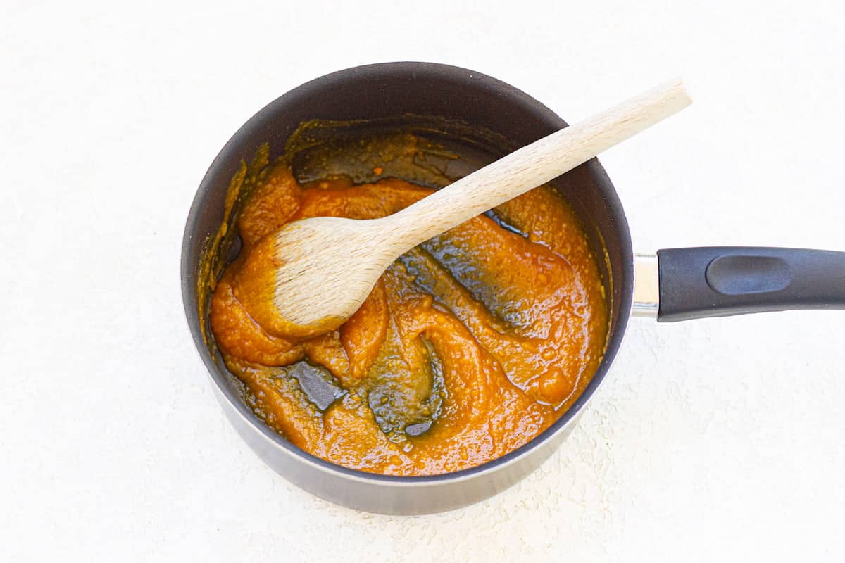 A saucepan with thick orange sauce being stirred by a wooden spoon.