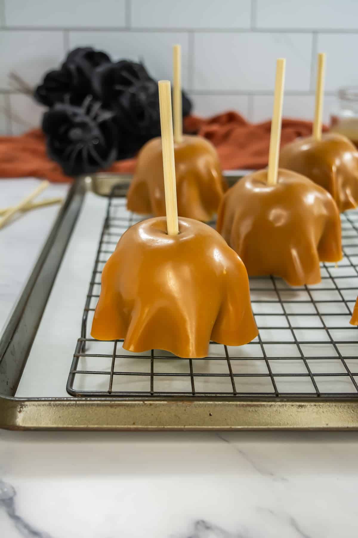 Caramel-covered treats with sticks on a cooling rack, set against a kitchen backdrop with a red cloth and black decorative items.