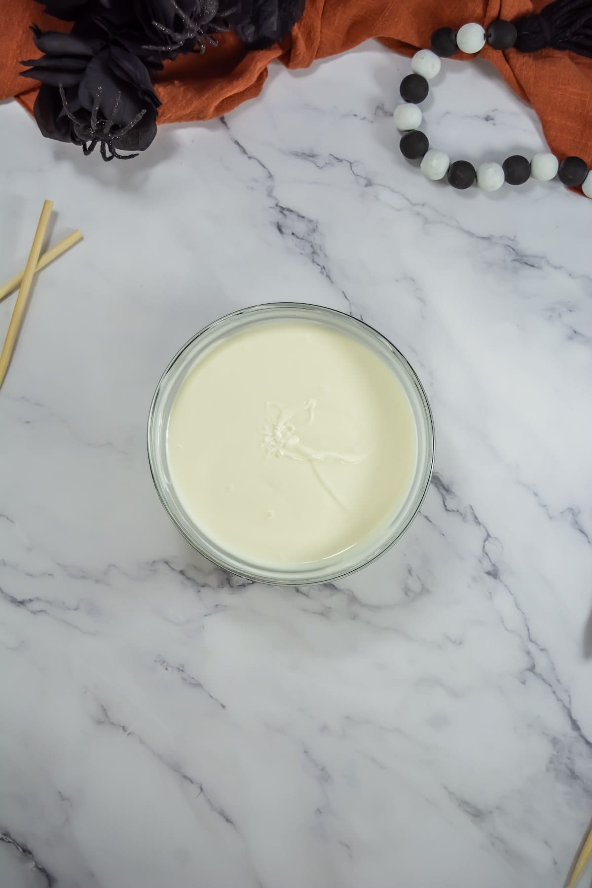A bowl of melted white chocolate on a marble surface, accompanied by two wooden sticks and a decorative black and white beaded item.