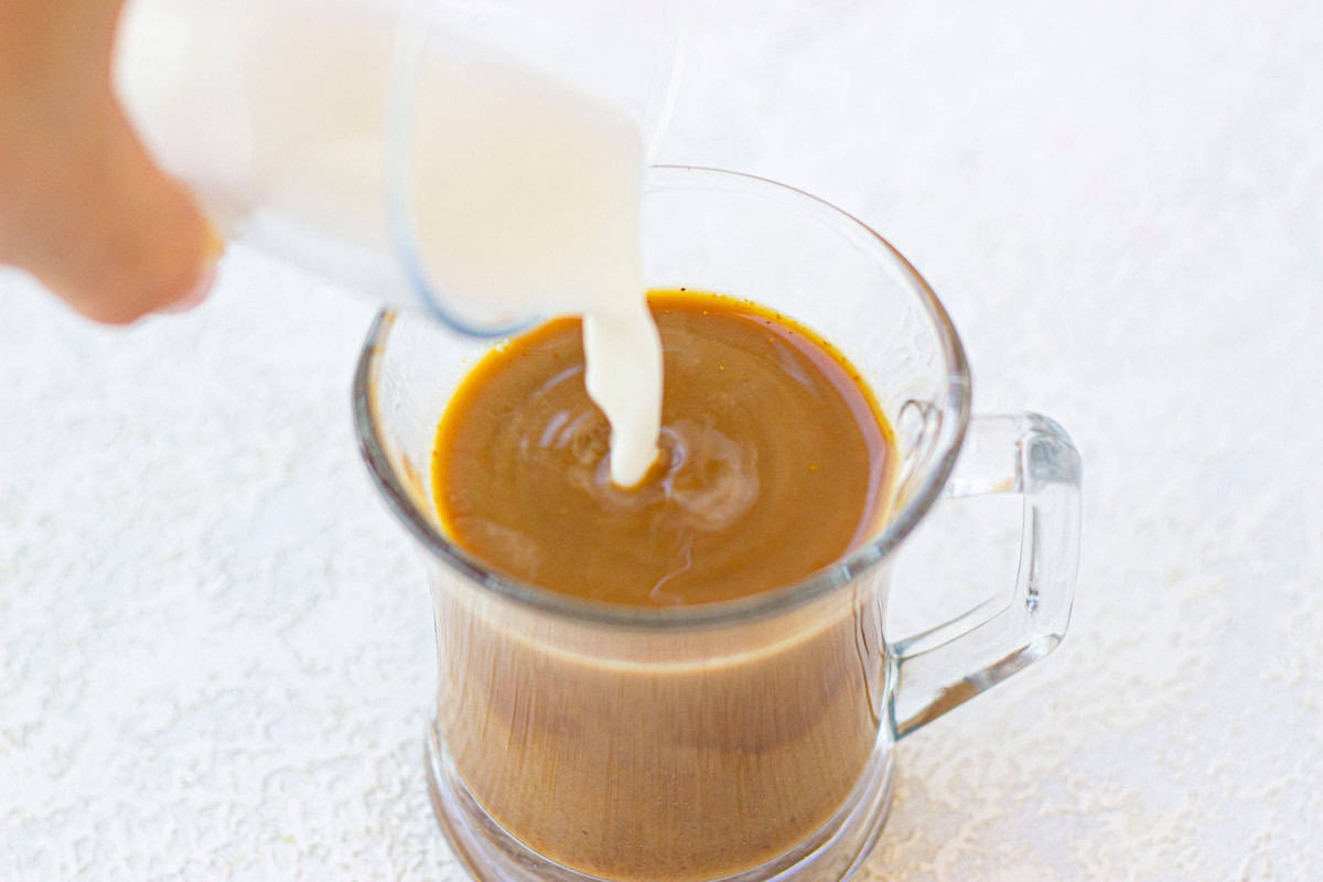 Milk being poured into a glass mug of coffee on a white surface.