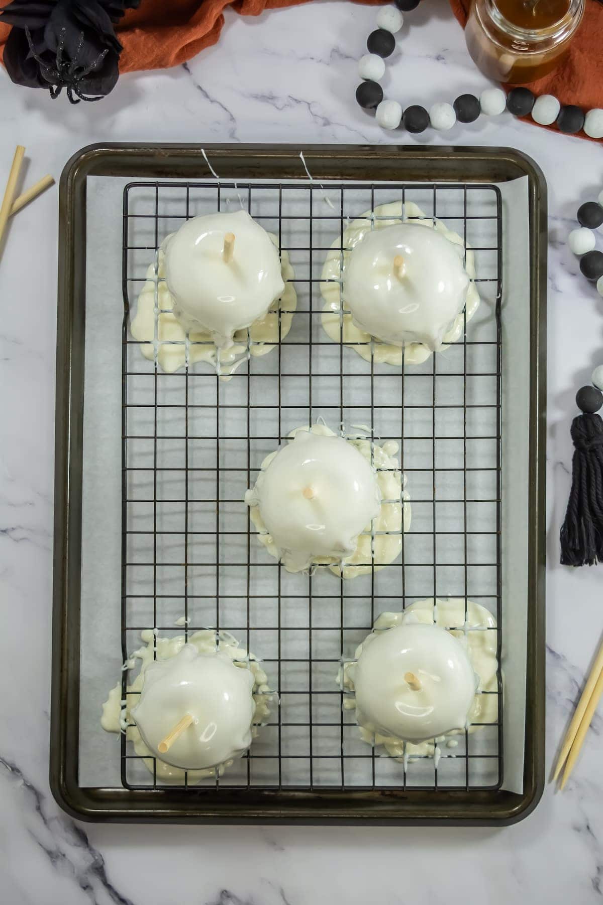 Five white chocolate-covered candy apples on a wire rack, each with a wooden stick, over a parchment-lined baking sheet on a marble surface.