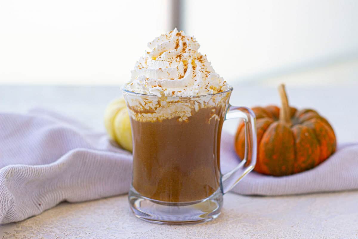 A glass mug of pumpkin spice latte topped with whipped cream and cinnamon. Two small pumpkins and a light cloth are in the background on a white surface.