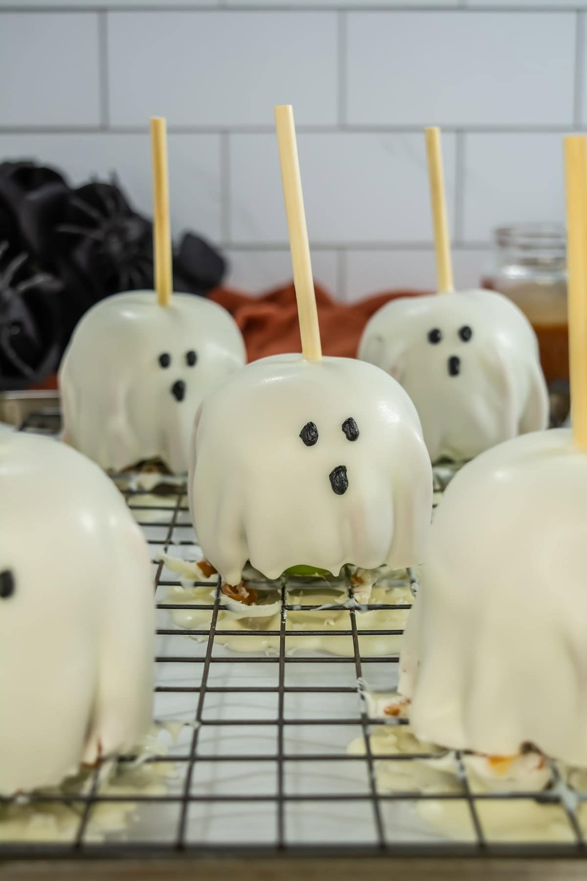 Five caramel apples covered in white chocolate, decorated with eyes and mouth, and skewered with sticks in the middle on top of a wire rack.
