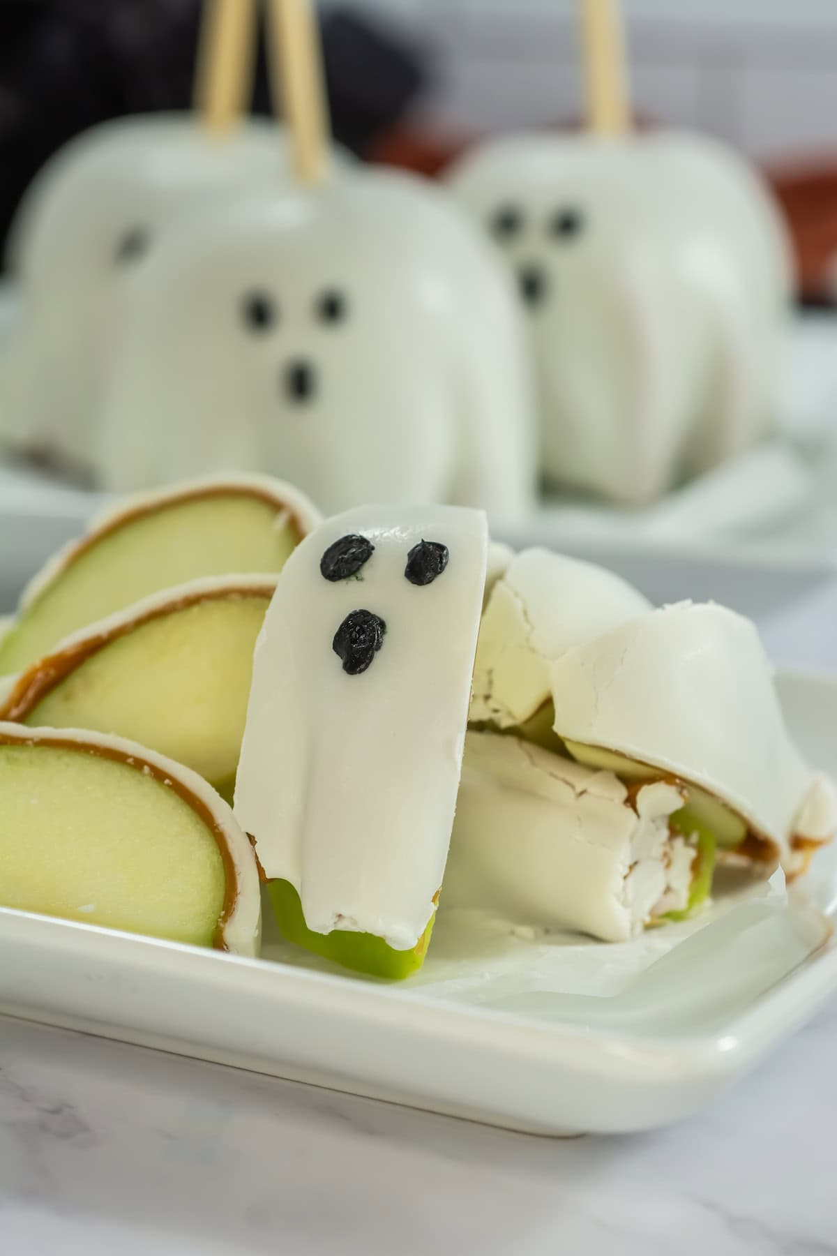 Sliced white chocolate-covered caramel apples on a plate in front, with three whole Halloween apples on the back.
