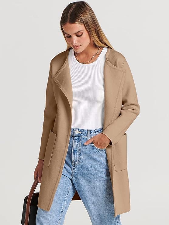 Person wearing a beige coat, white ribbed top, and blue jeans, holding a bag, standing against a plain background.