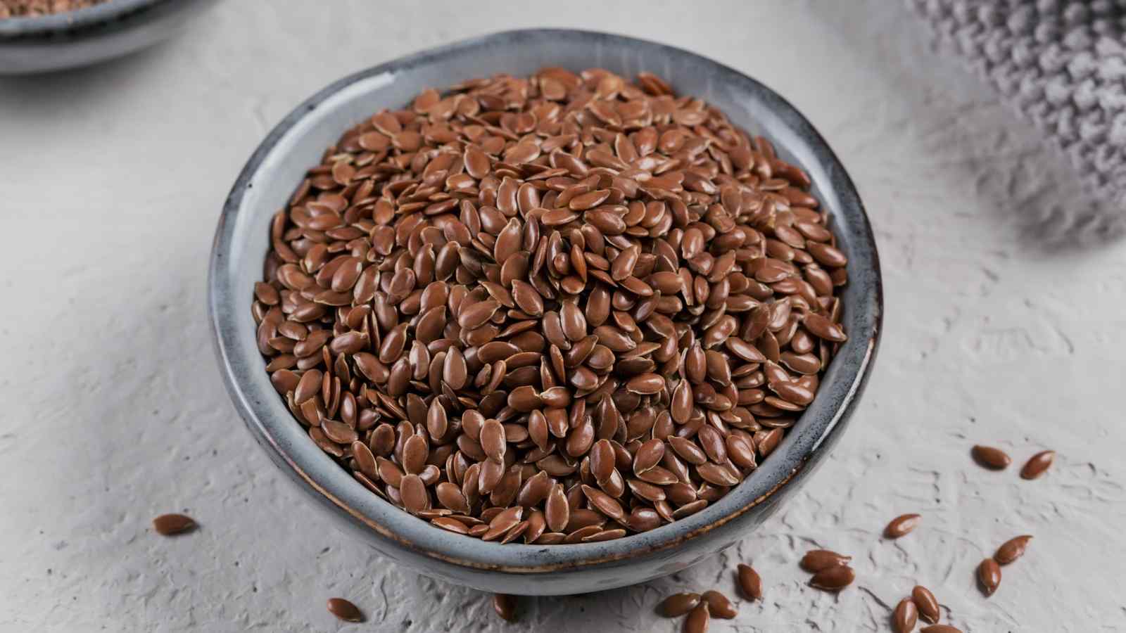 A bowl filled with brown flaxseeds on a light gray surface.