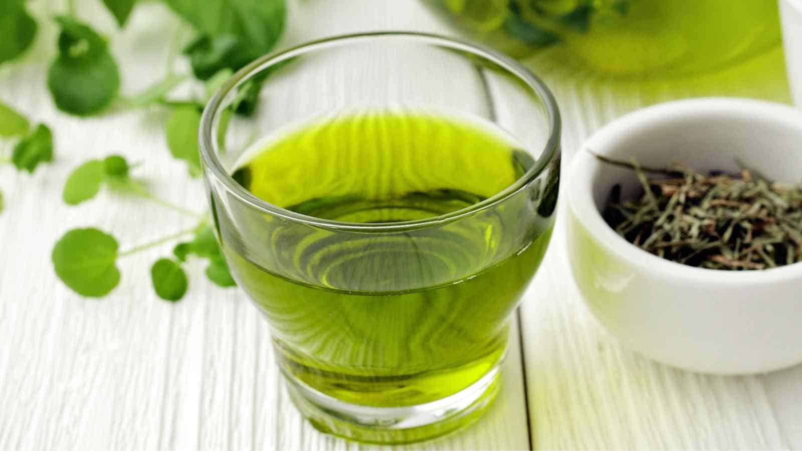 A glass of green liquid placed on a white wooden table next to a small white bowl containing dried herbs.
