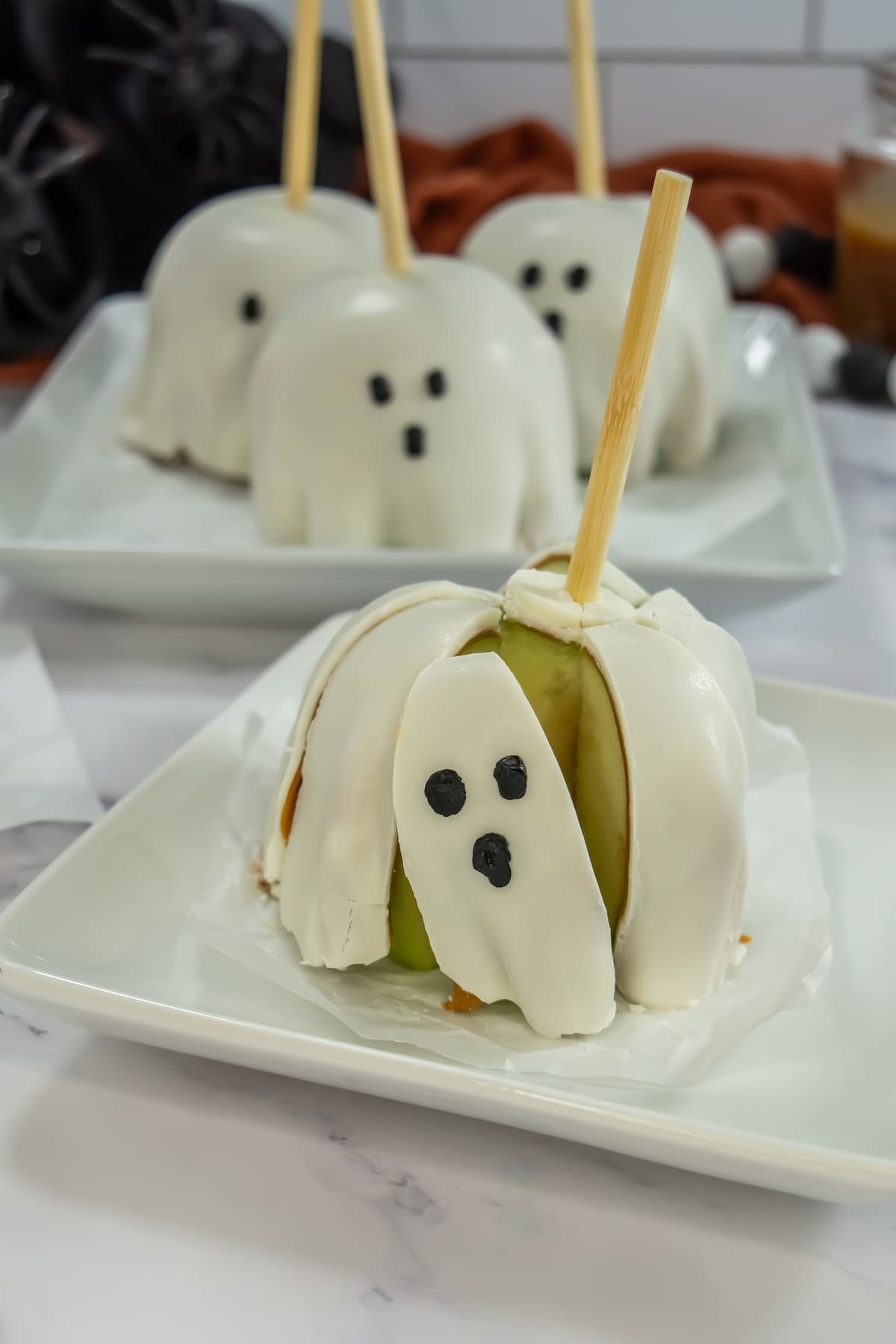 A sliced Halloween caramel apple decorated with eyes and mouth on a white square plate. Behind it are three whole white-chocolate covered apples, also on a square plate.