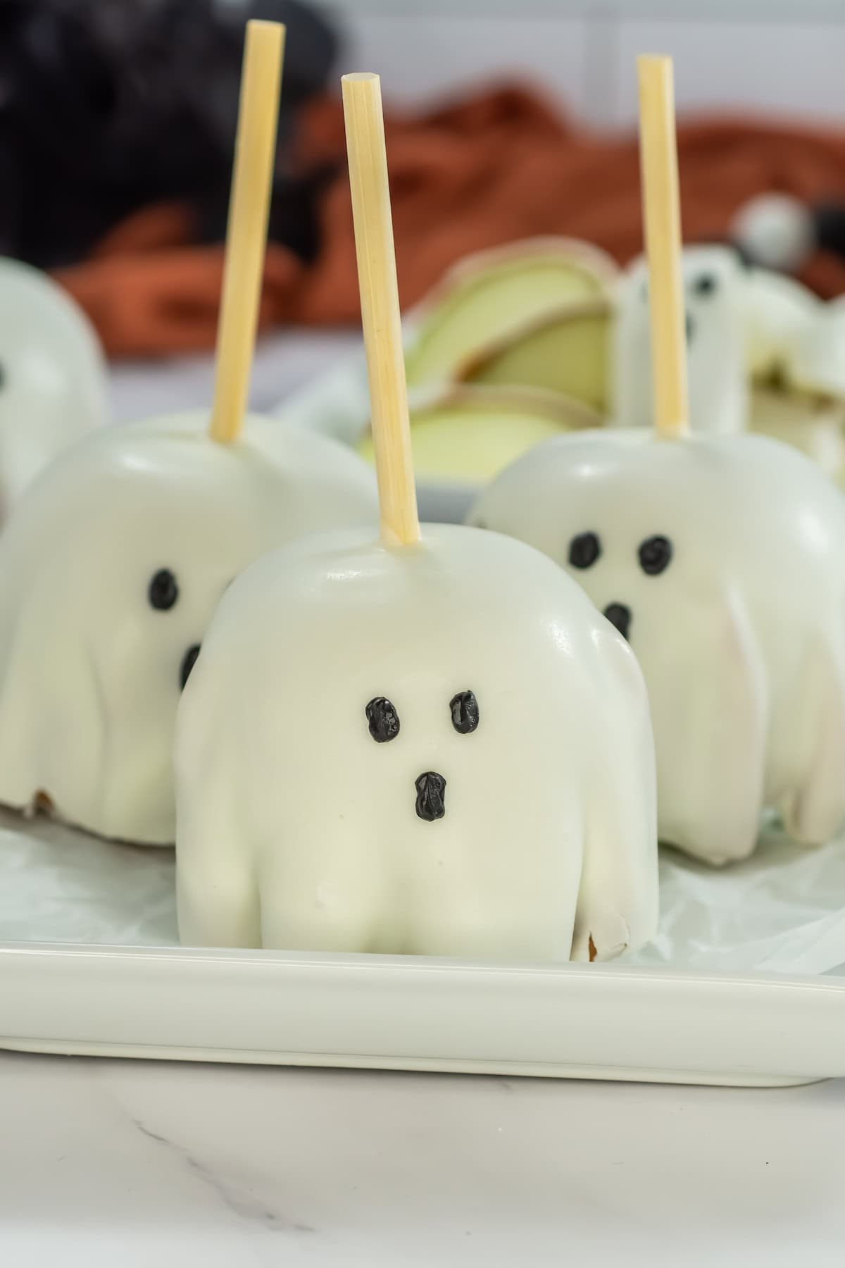 Three white chocolate-covered caramel apples on a square white plate, each decorated with eyes and mouth and skewered with a wooden stick.
