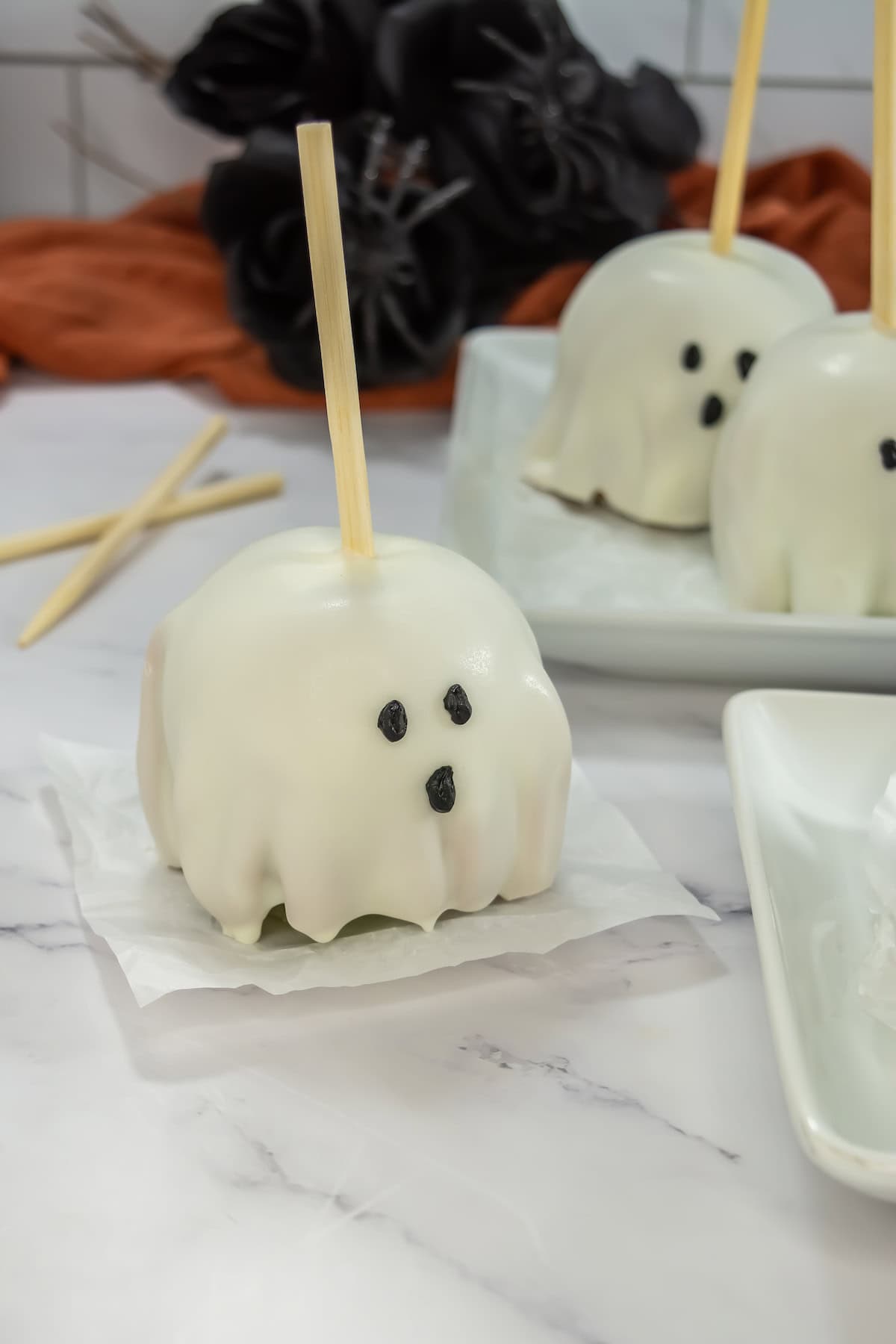 A white-chocolate covered apple decorated with eyes and mouth on top of a square parchment paper. Behind it are two white-colored apples on a square white plate.
