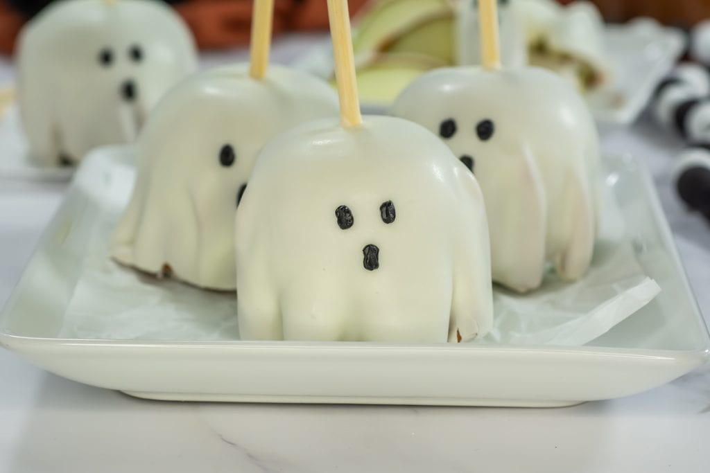 Three white-covered caramel apples on a square white plate, each decorated with eyes and mouth and skewered with a wooden stick in the middle. Behind the plate is another white apple on parchment paper.