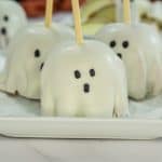 Three white-covered caramel apples on a square white plate, each decorated with eyes and mouth and skewered with a wooden stick in the middle. Behind the plate is another white apple on parchment paper.