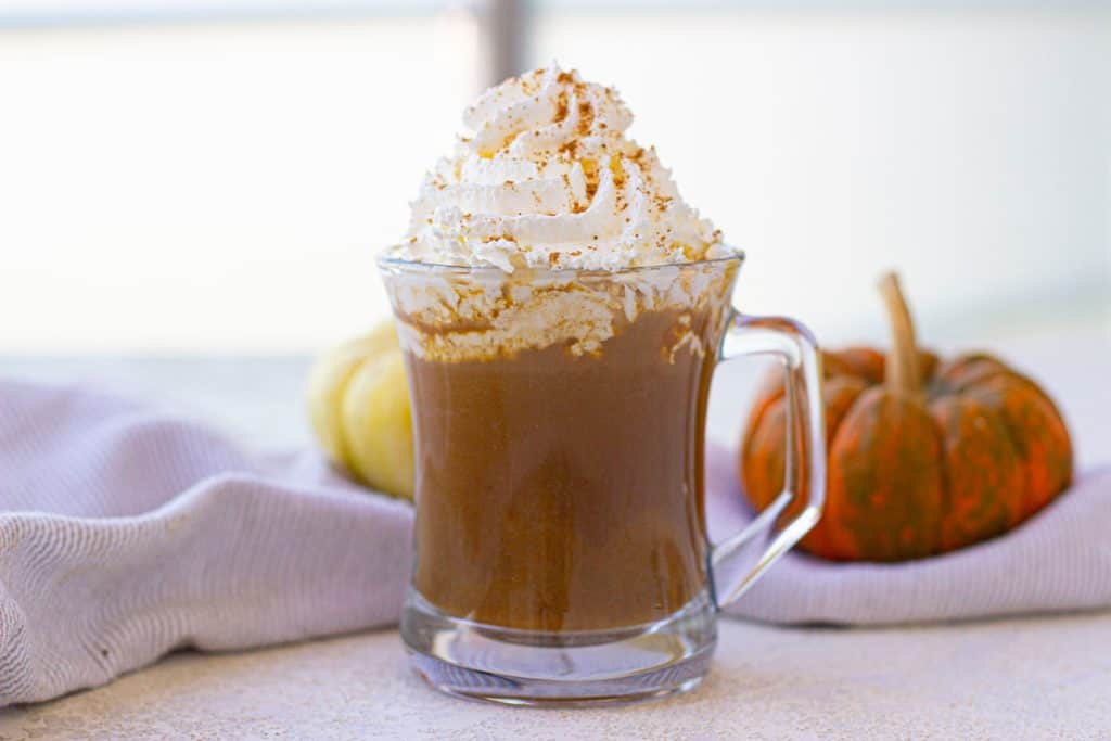 A glass mug of pumpkin spice latte topped with whipped cream and cinnamon, set on a table with a white cloth. Two small pumpkins are in the background.