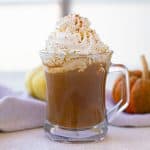 A glass mug of pumpkin spice latte topped with whipped cream and cinnamon, set on a table with a white cloth. Two small pumpkins are in the background.