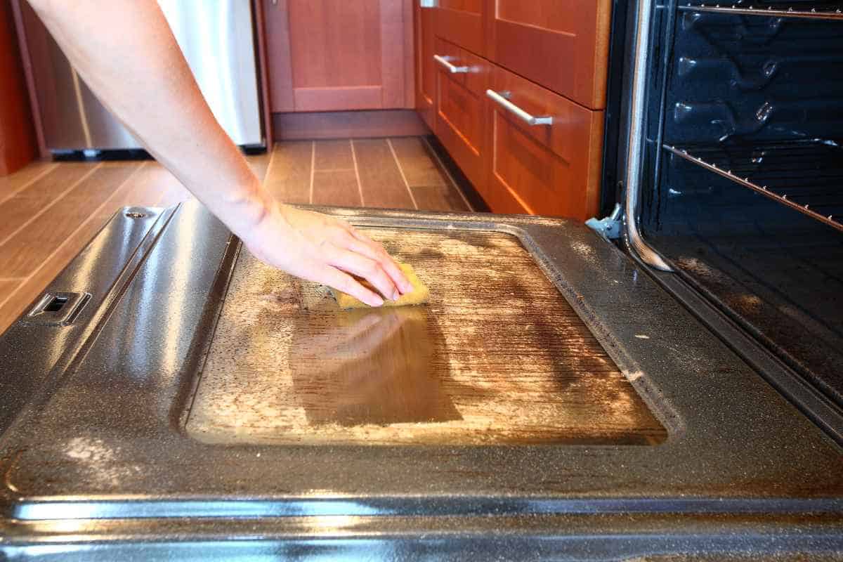 A person demonstrates how to clean an oven efficiently by using a sponge on the open door in a tidy kitchen setting.