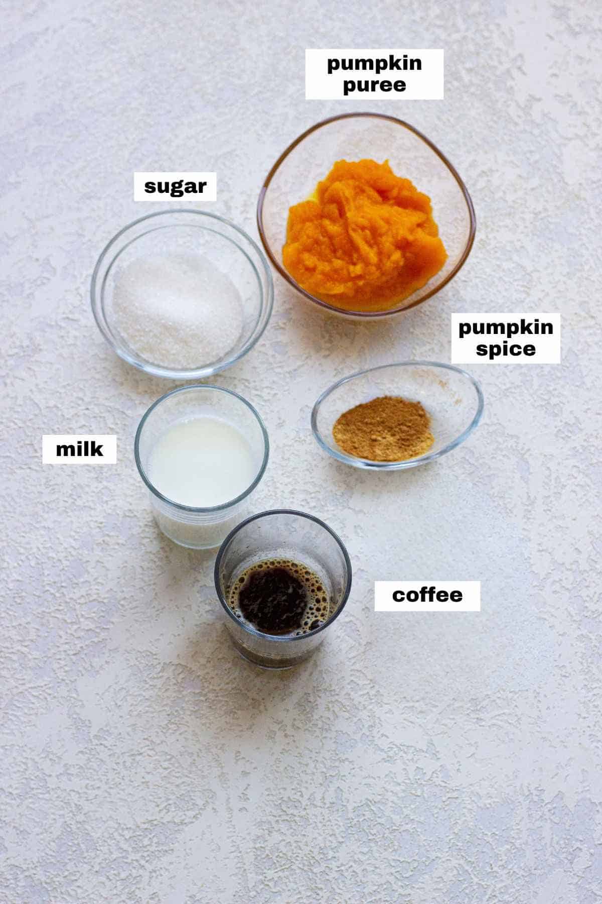 Ingredients for a pumpkin spice latte arranged on a surface: pumpkin puree, sugar, pumpkin spice, milk, and a shot of coffee.