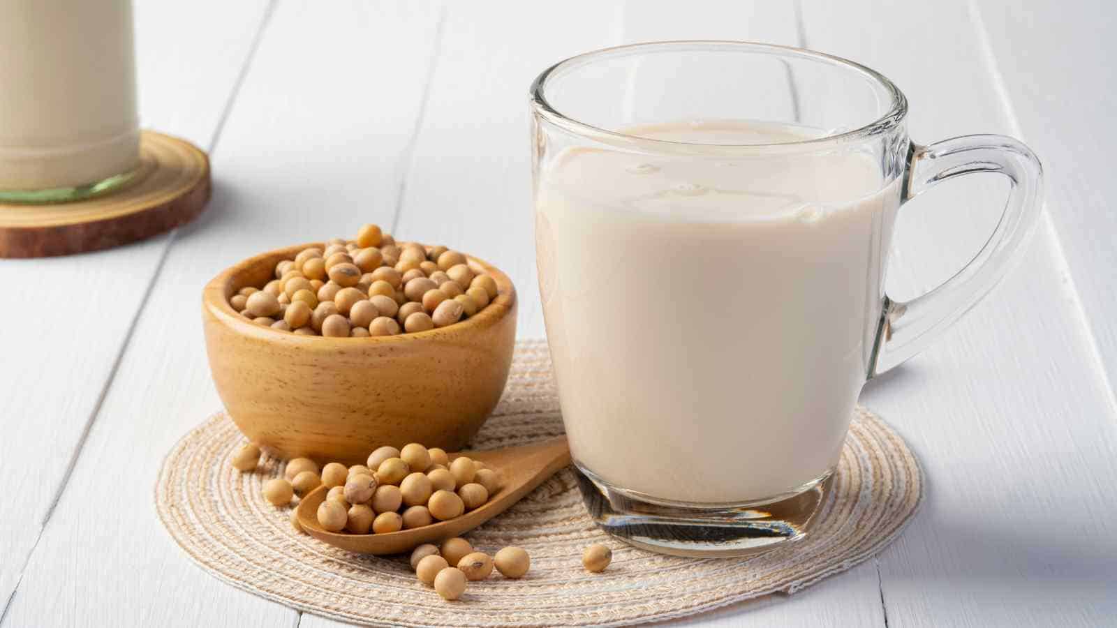A glass of soy milk sits on a woven mat beside a wooden bowl and spoon filled with soybeans, on a white surface.