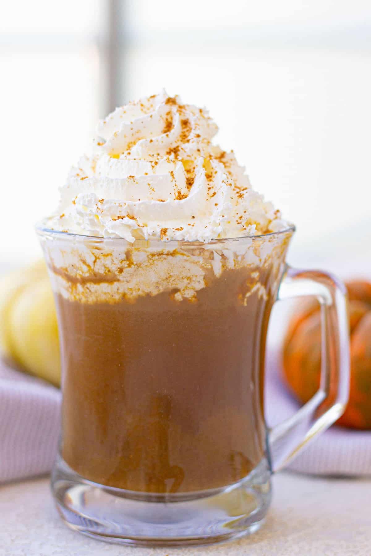 Glass mug of hot pumpkin spice latte topped with whipped cream and a sprinkle of cinnamon, with blurred pumpkins in the background.