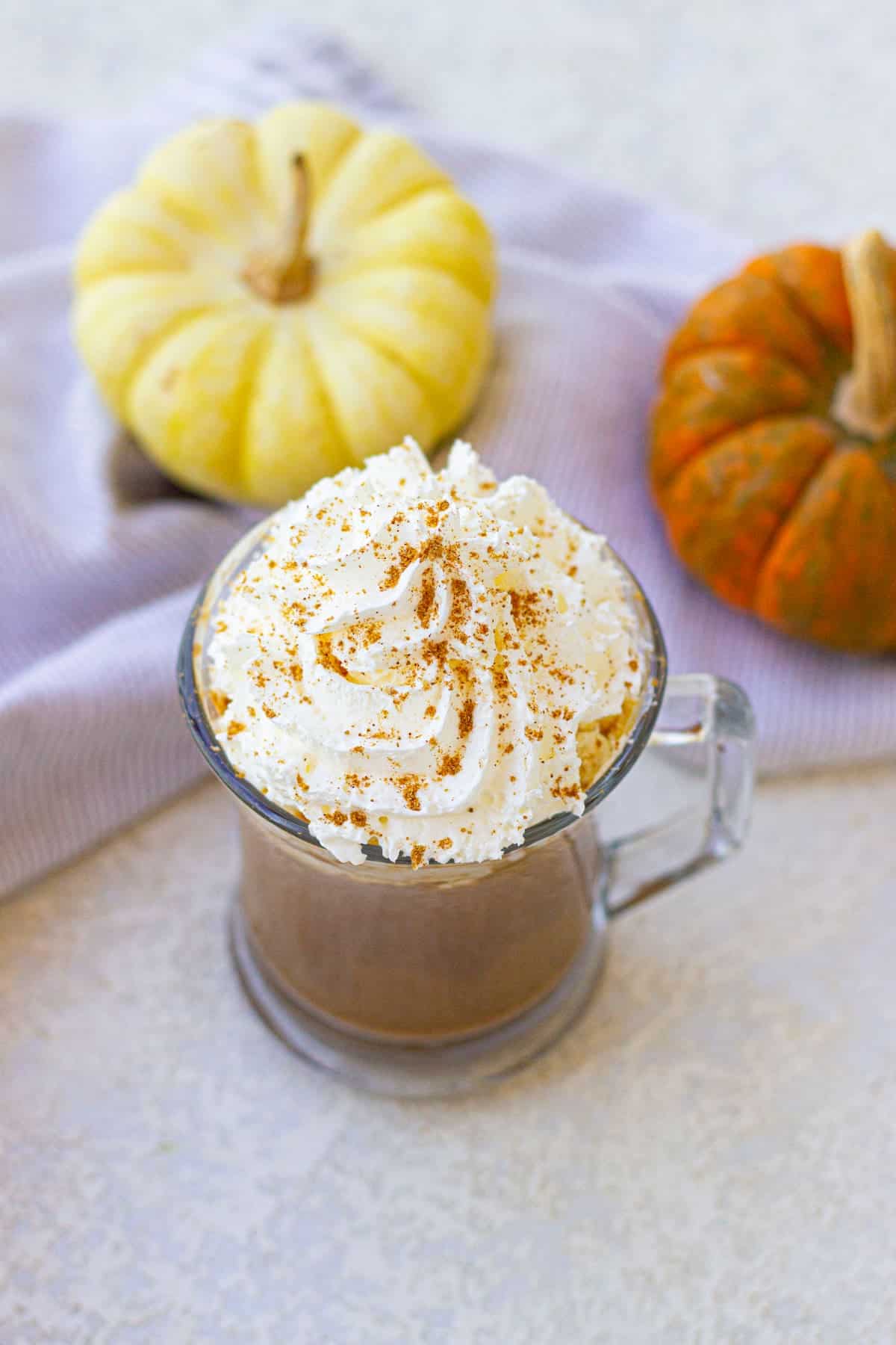 A mug of whipped cream-topped pumpkin spice latte with a dusting of spices, set on a cloth napkin, accompanied by two small pumpkins, one yellow and one orange.