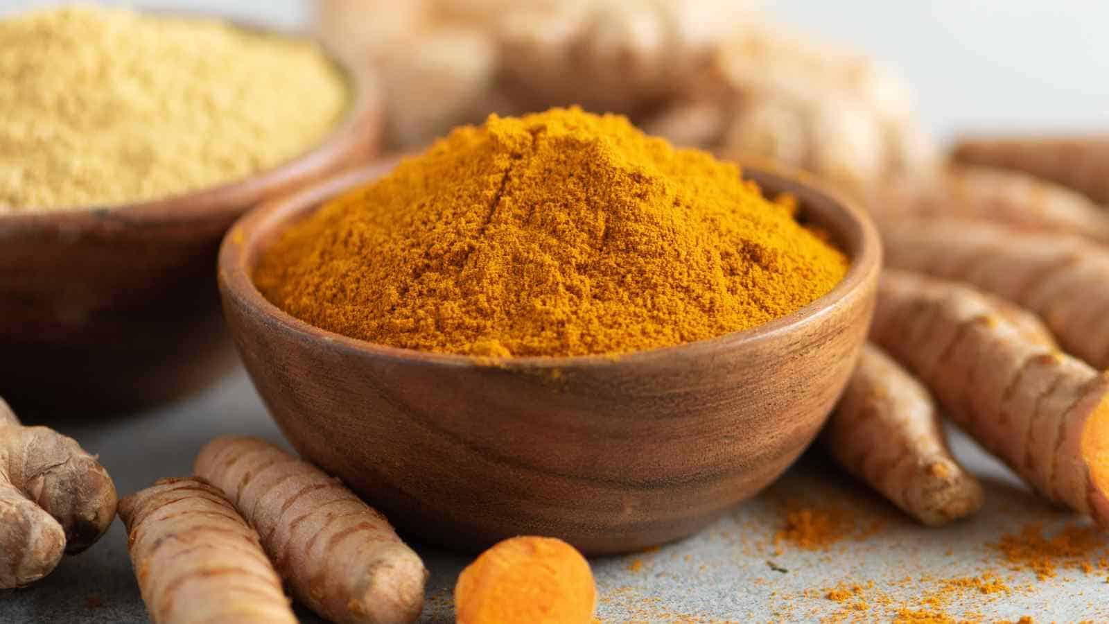 A wooden bowl filled with ground turmeric is surrounded by fresh turmeric roots on a table.