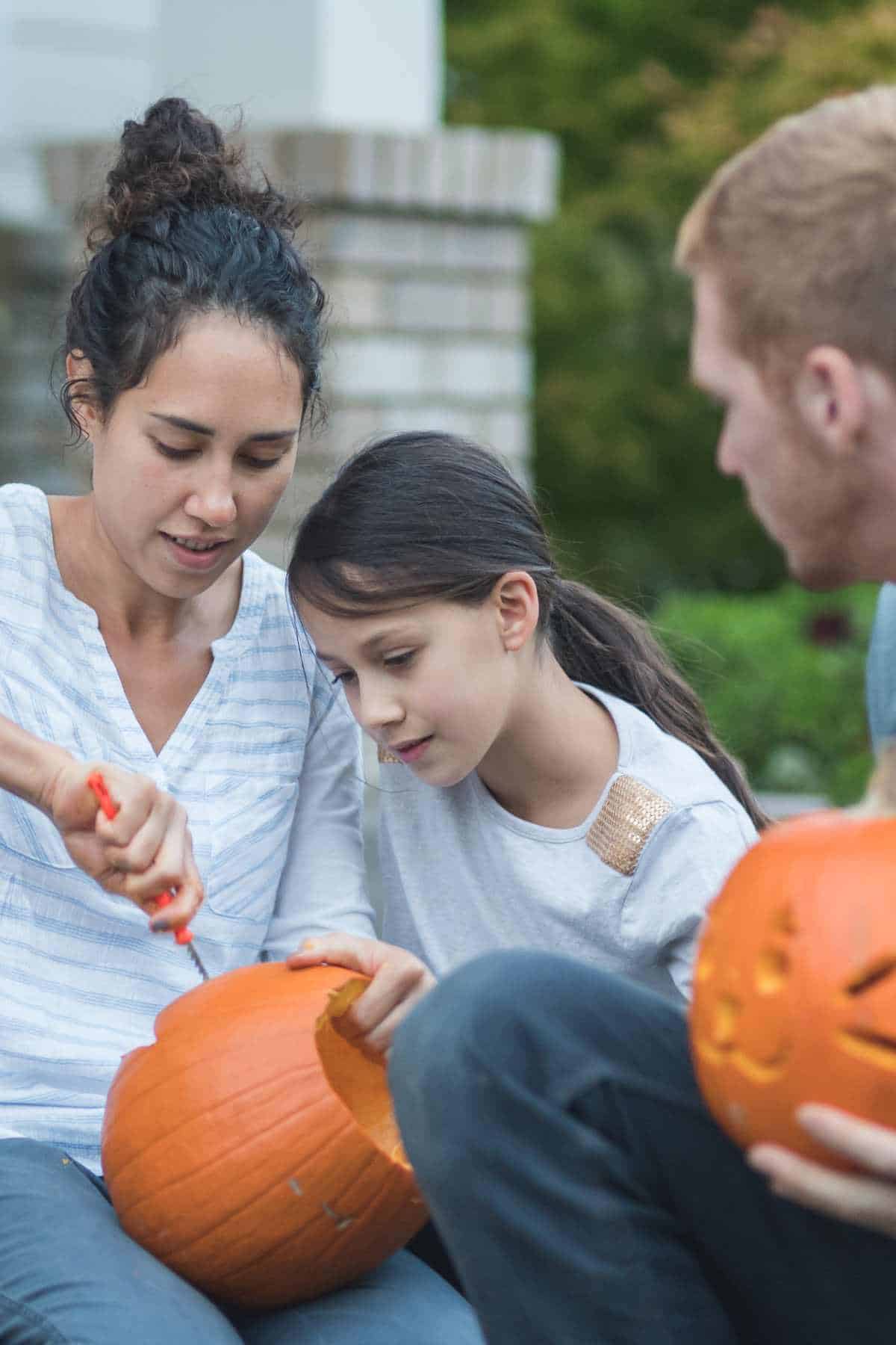 Two adults and a child are seated outdoors, carving pumpkins. One of the adults is actively carving while the child watches closely. Another carved pumpkin is visible in the foreground.