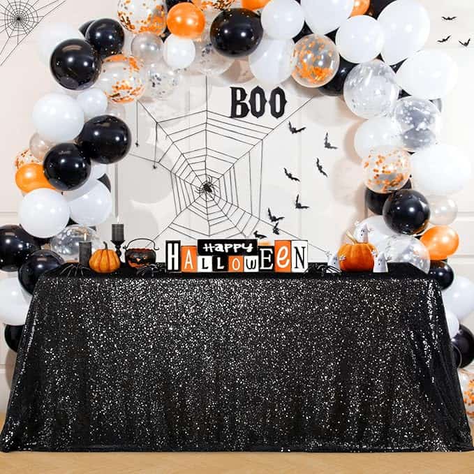 A decorated Halloween table with black sequin cloth, balloon arch in orange, black, and white, and a "Happy Halloween" sign.