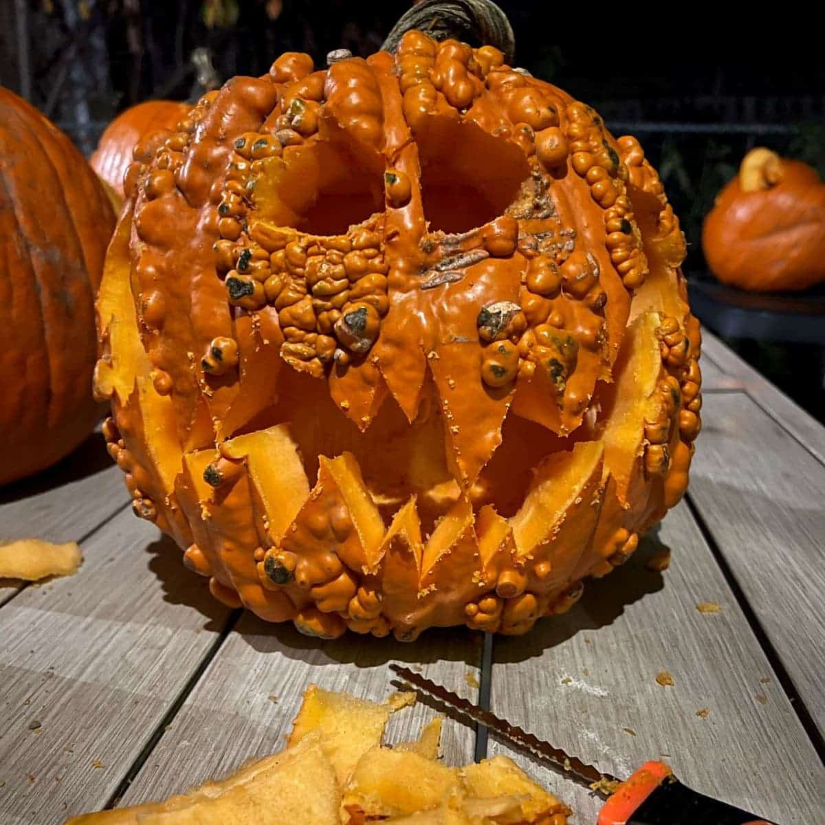 A carved, bumpy pumpkin with sharp teeth and eyes sits on a wooden table next to carving tools.