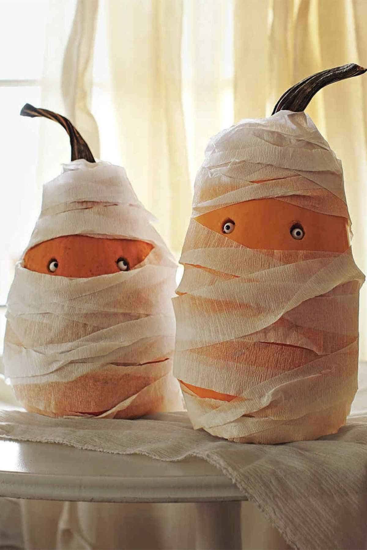 Two pumpkins wrapped in white gauze with painted eyes, resembling mummies, are displayed on a round table.