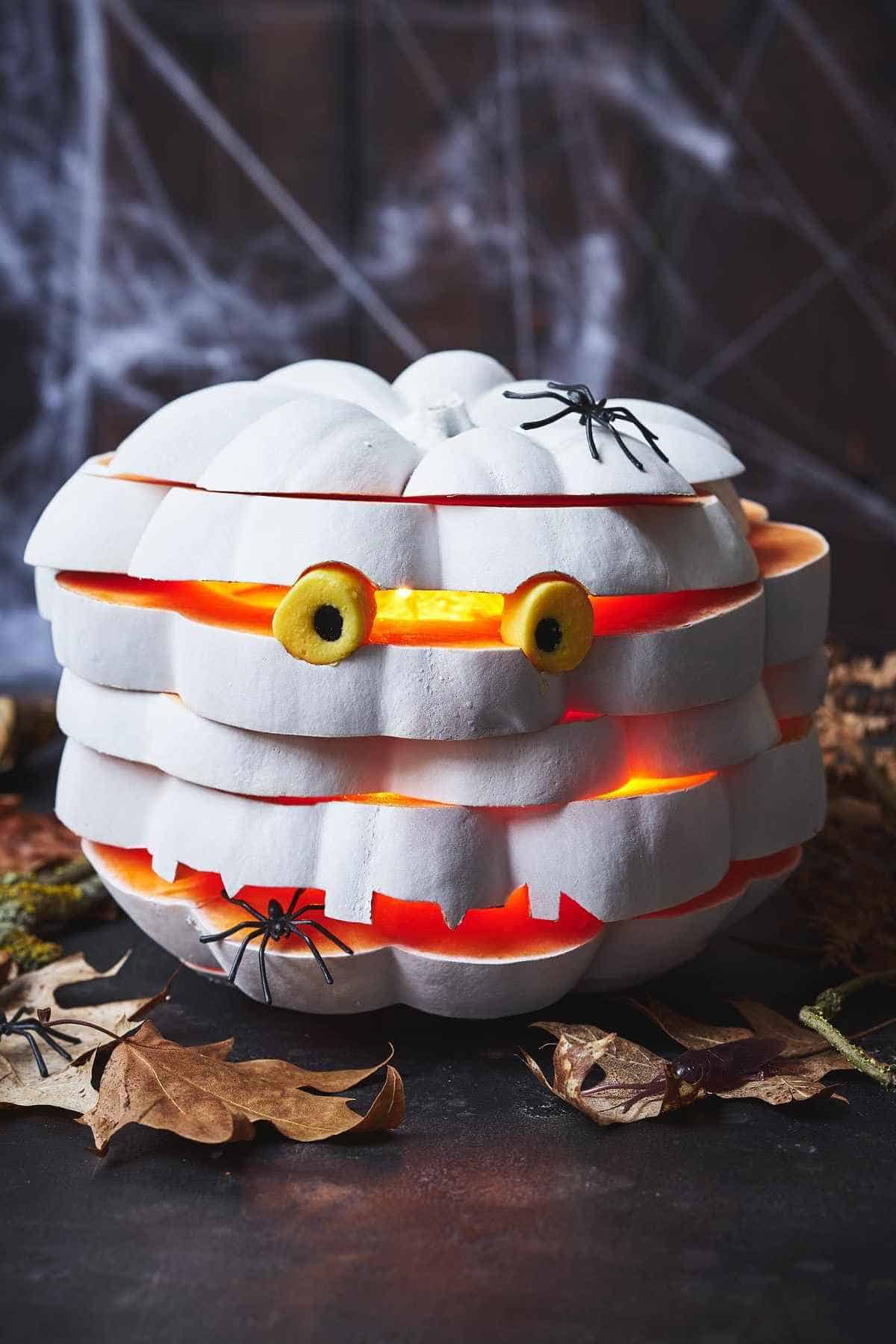 A white pumpkin carved in a layered style with glowing eyes, surrounded by artificial spiders and dried leaves.