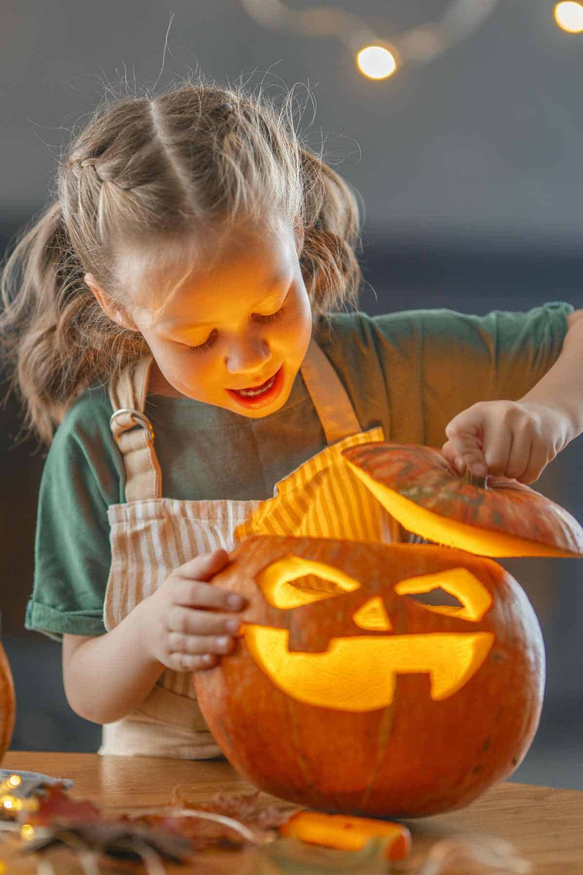 A young girl, wearing a striped apron, lifts the top off a carved, lit jack-o'-lantern, smiling with excitement.