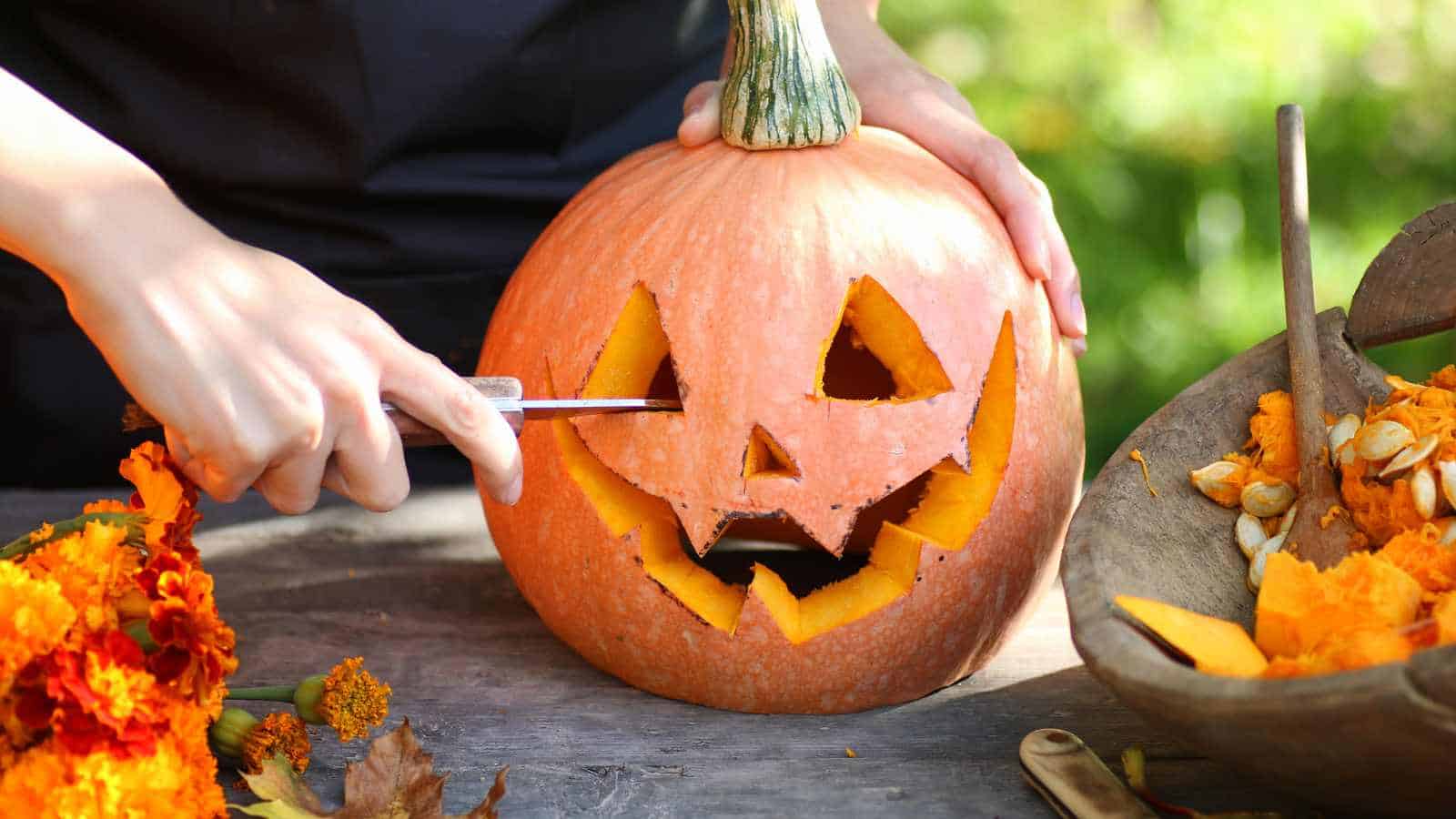 Person carving a jack-o'-lantern face into a pumpkin with a knife, surrounded by pumpkin seeds and flowers on a wooden table.
