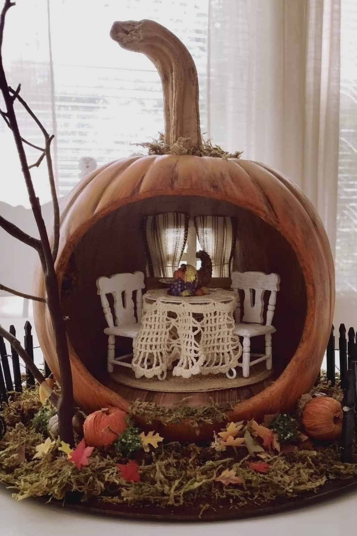 A large hollow pumpkin is carved to resemble a miniature room with two chairs and a table inside, surrounded by small pumpkins and autumn leaves.