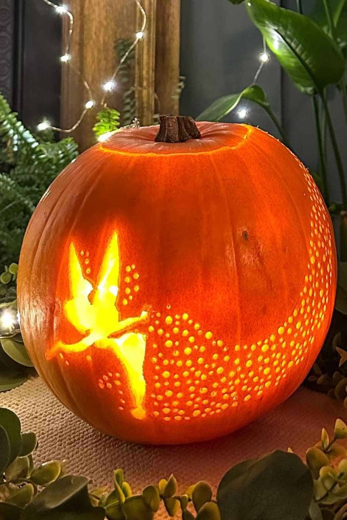 Carved pumpkin with a Tinker Bell design glowing in the dark, surrounded by plants and fairy lights.