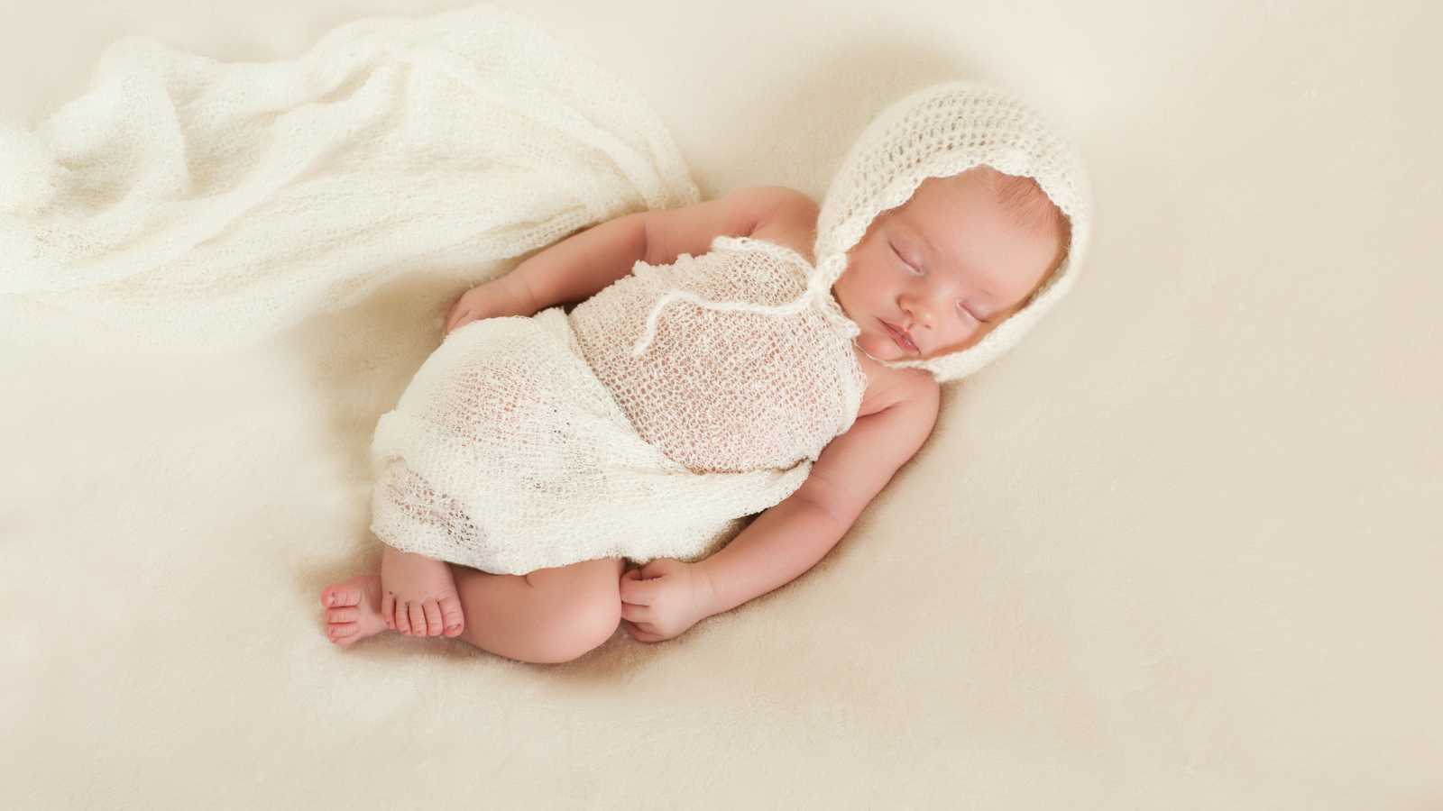 A sleeping baby wrapped in a white knitted blanket and bonnet lies on a cream-colored surface.