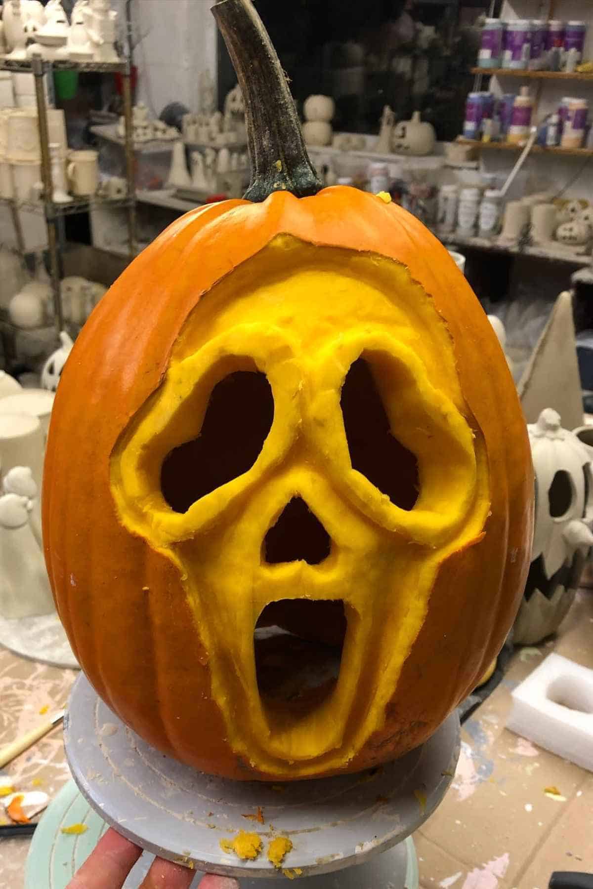 A pumpkin carved with a scream-like face sits on a table surrounded by pottery and craft supplies.