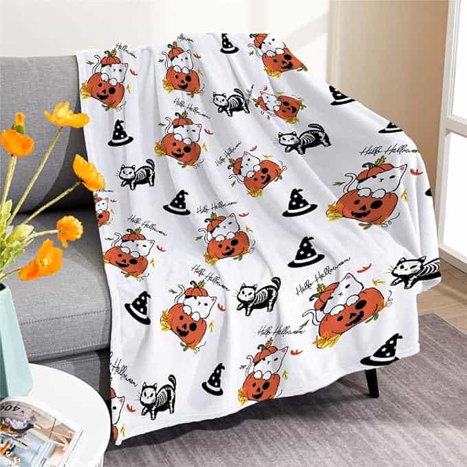 White Halloween-themed blanket on a couch with pumpkin and cat designs, witch hats, and "Happy Halloween" text.