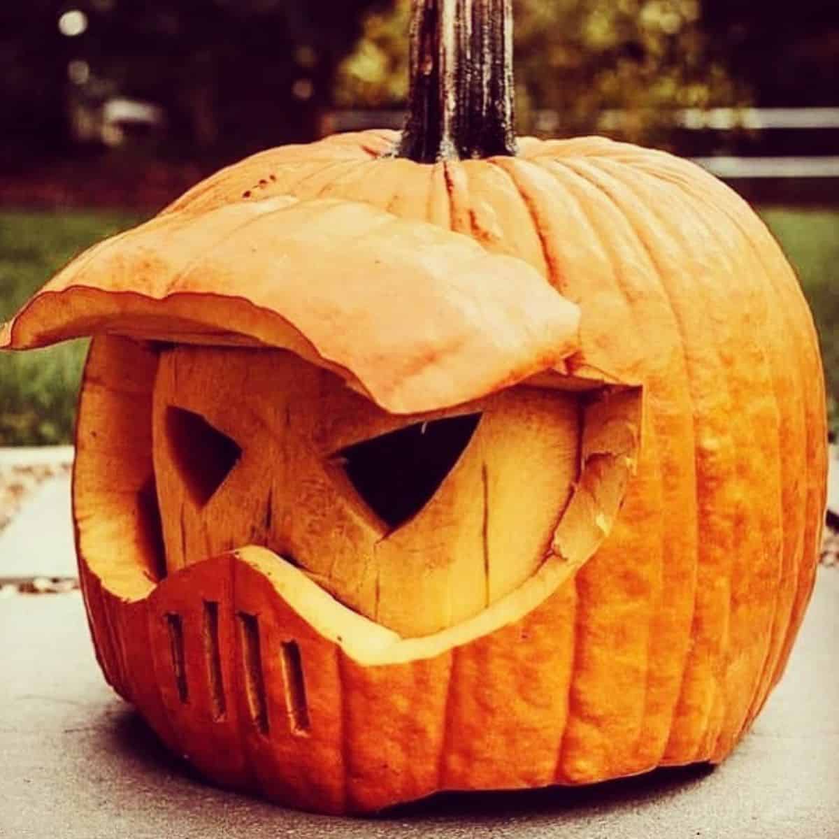 Carved pumpkin with a menacing face, featuring a visor-like design and a grill pattern below the eyes, placed outdoors on a gray surface.