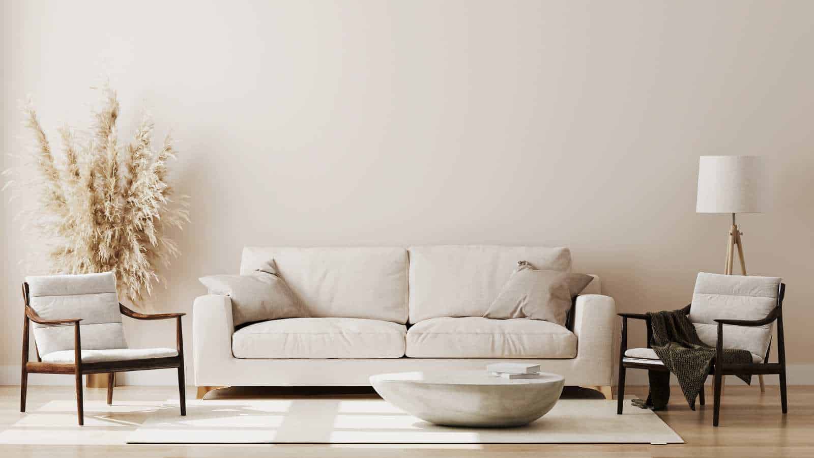 Minimalist living room with a beige sofa, two chairs, a round coffee table, a floor lamp, and a tall vase of pampas grass against a light wall.