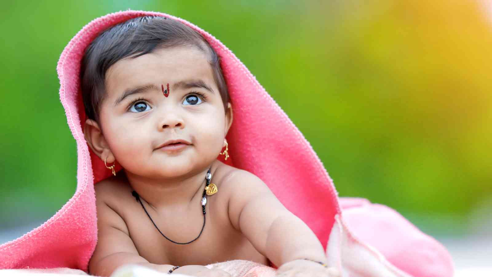 A baby with a pink blanket draped over their head, wearing a small bindi, necklace, and earrings, gazes upward against a blurred green and yellow background.