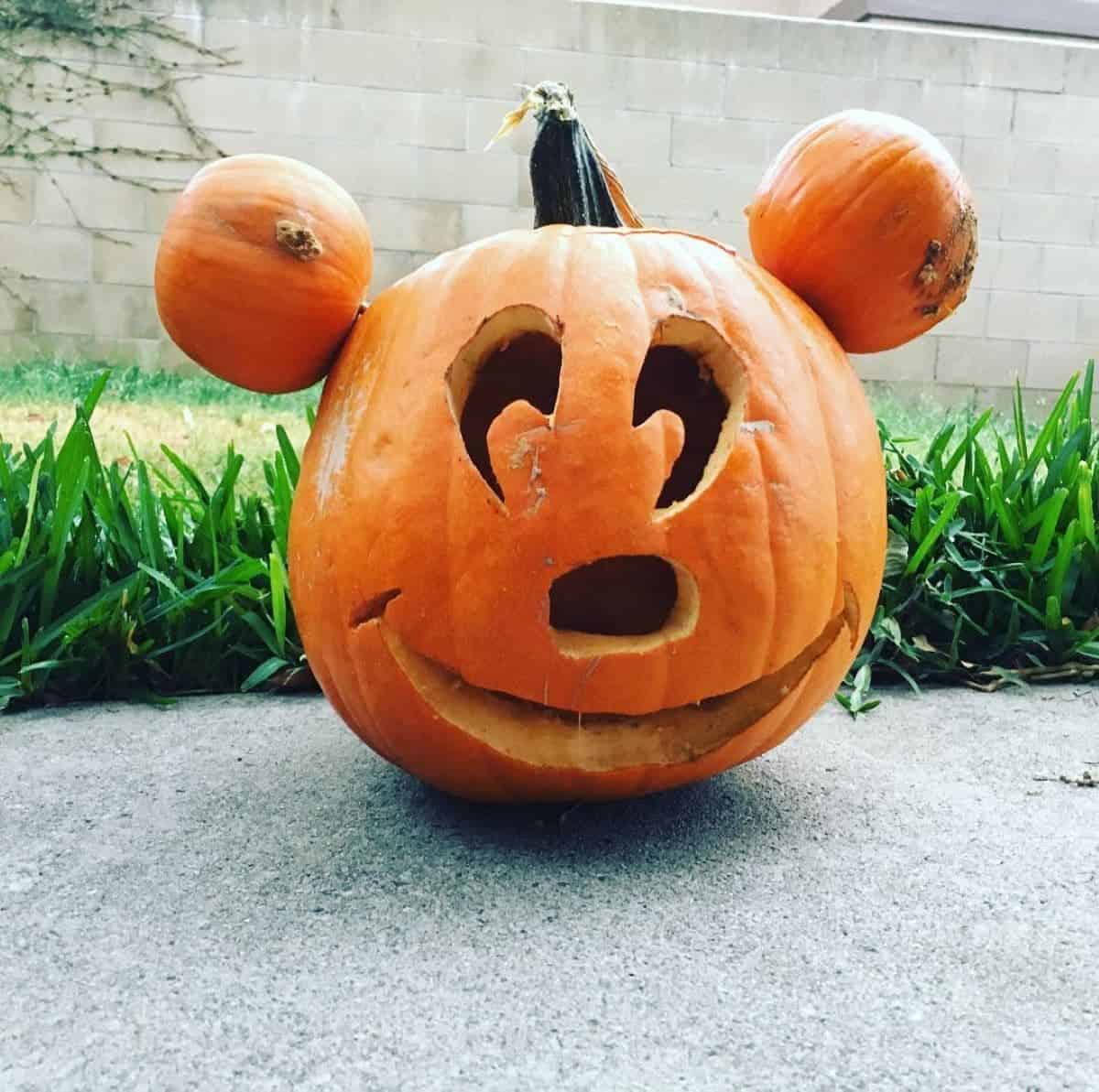 A carved pumpkin resembling a cartoon character with two smaller pumpkins as ears, placed on a concrete surface with grass and a brick wall in the background.