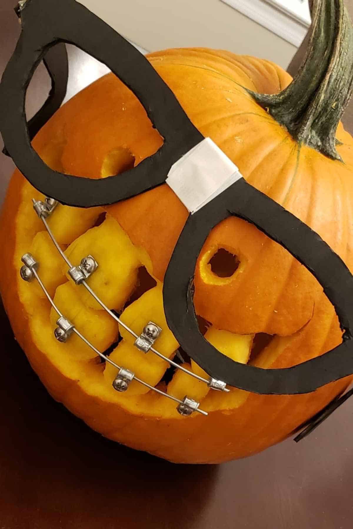 A carved pumpkin with glasses and braces made from metal wires, featuring a crooked smile.