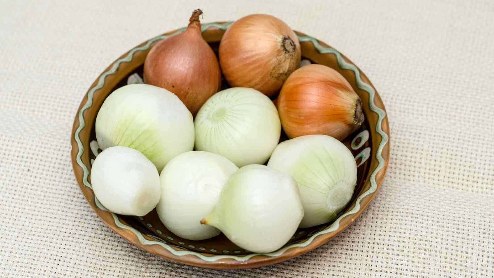 A bowl containing a mix of white and brown onions on a textured surface.