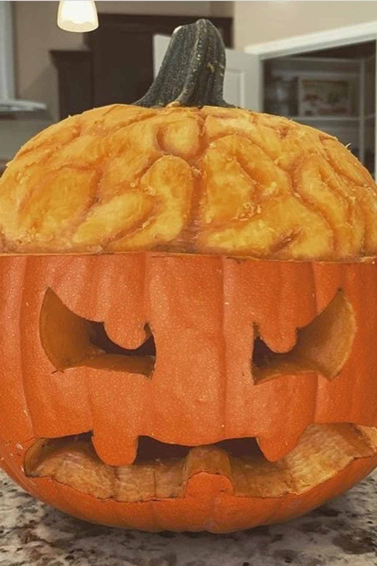 Carved pumpkin with an expressive face, featuring cut-out eyes, nose, and mouth, placed on a textured surface indoors.