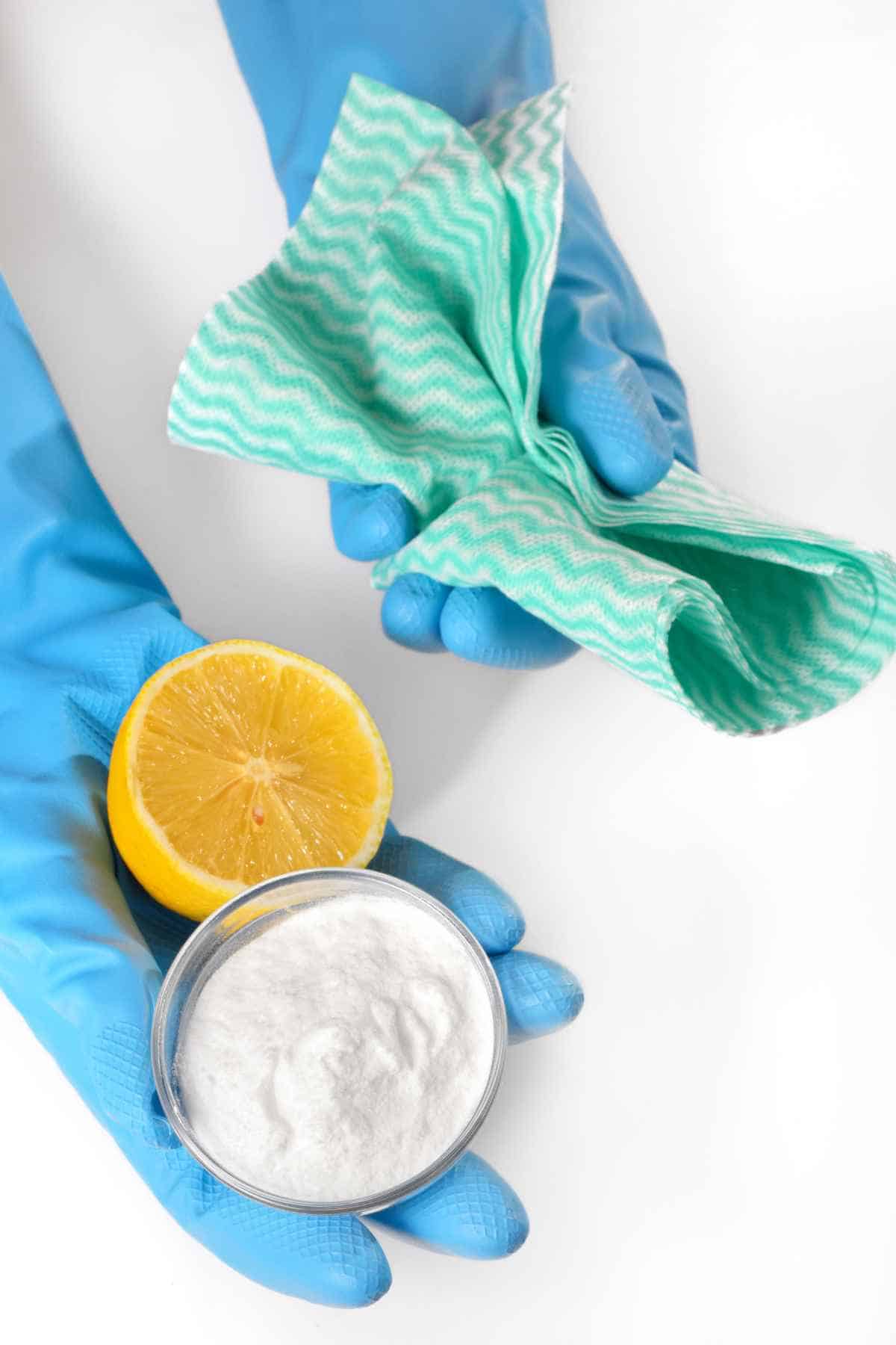A person wearing blue rubber gloves holding a half of a lemon, a small bowl of baking soda and a green cleaning cloth.