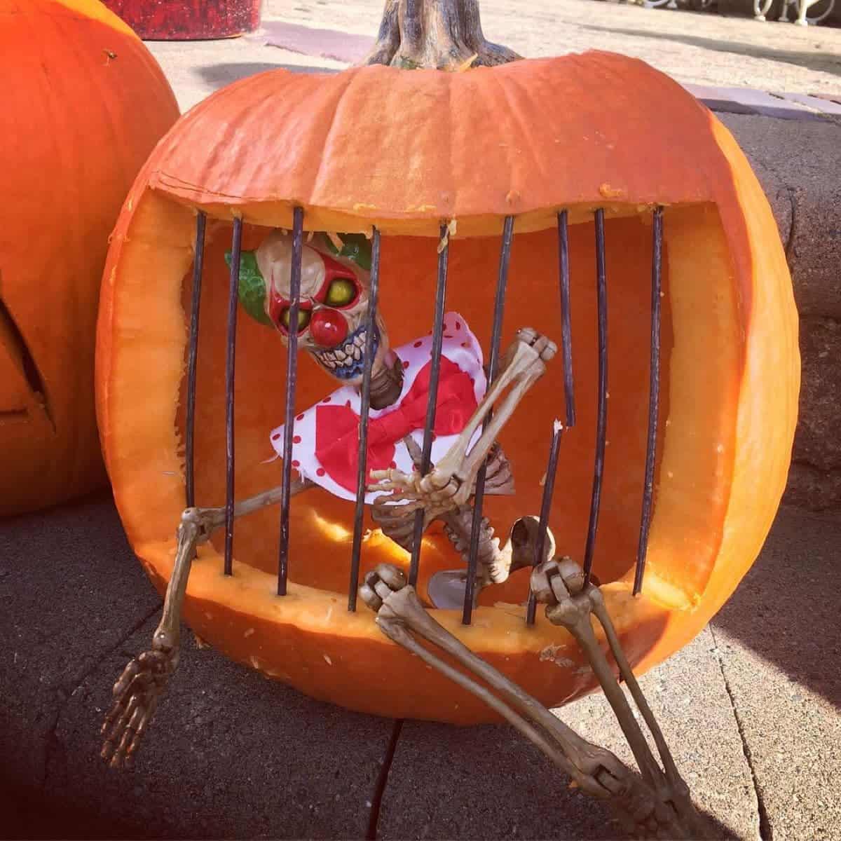 Pumpkin carved to resemble a cage, containing a skeleton with clown features, positioned on a concrete surface.