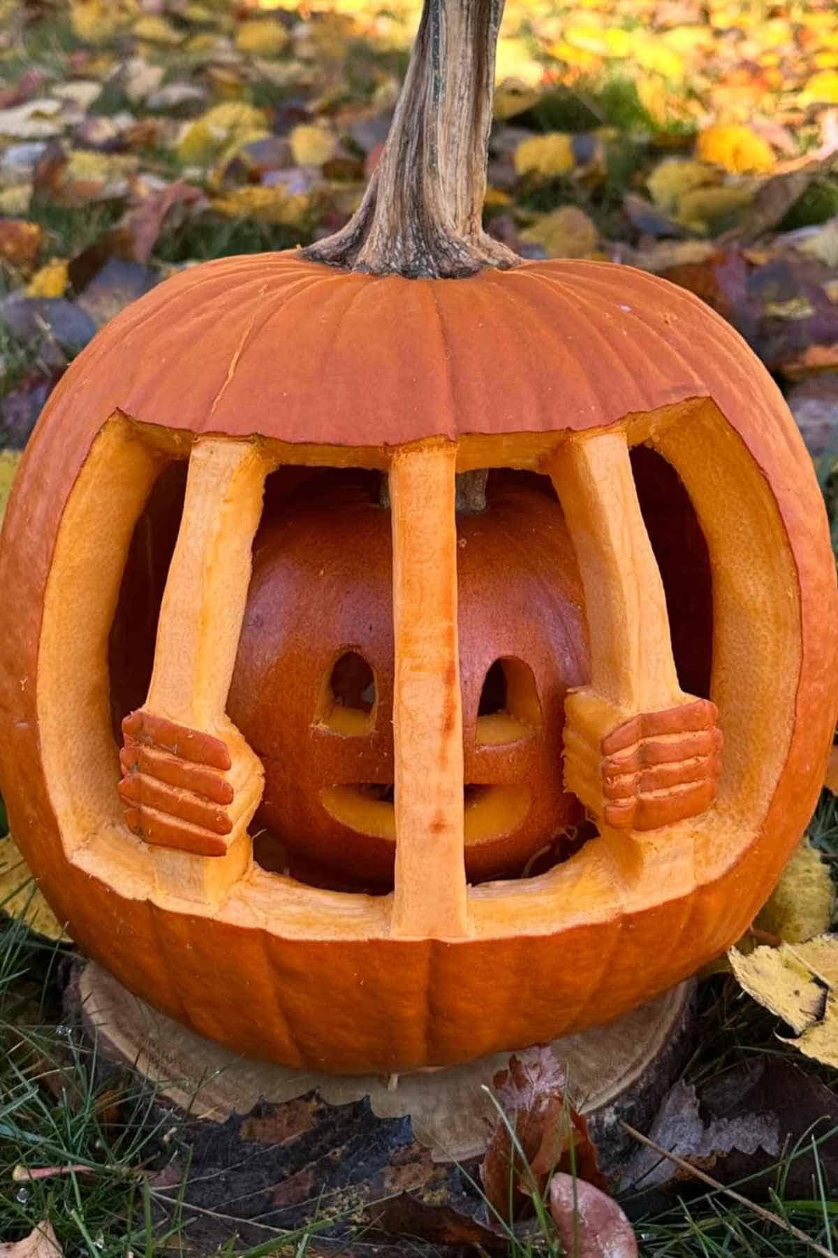 A carved pumpkin showing a smaller pumpkin face behind pumpkin bars, resembling a jail cell, on a grassy area with autumn leaves.