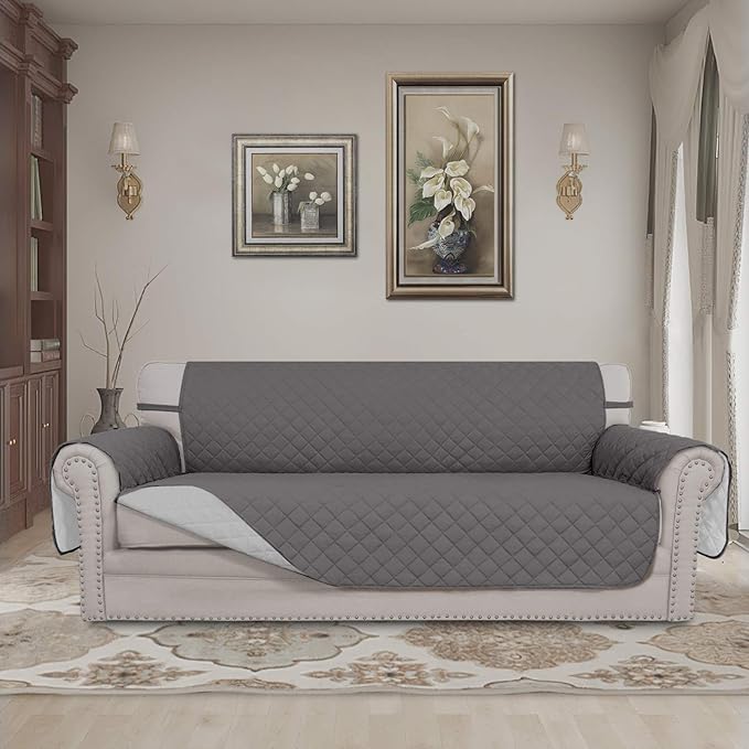 A gray quilted sofa cover on a light-colored sofa in a living room with framed floral artwork, two wall lamps, and a decorative rug.