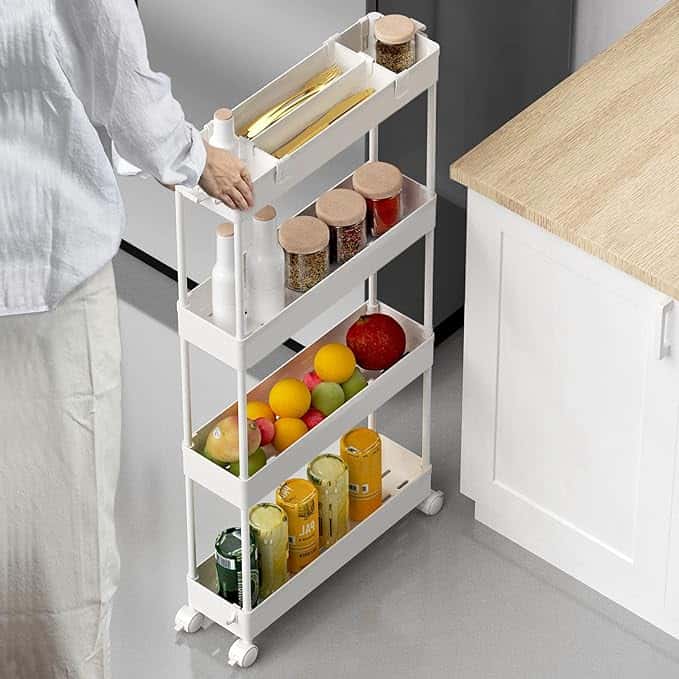 A person arranges items on a narrow, four-tier white rolling cart filled with jars, bottles, and fruit in a kitchen.