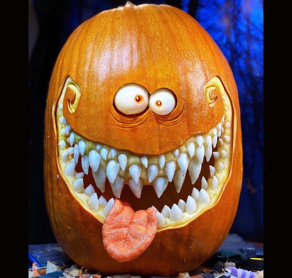 A carved pumpkin with a large, toothy grin and protruding eyes and tongue, set against a dark blue background.