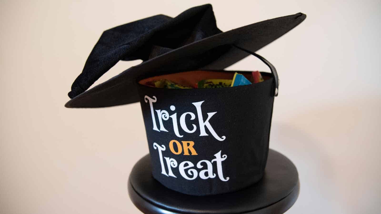 A black bucket with "Trick or Treat" text is topped with a witch's hat, sitting on a stool, and filled with candy.